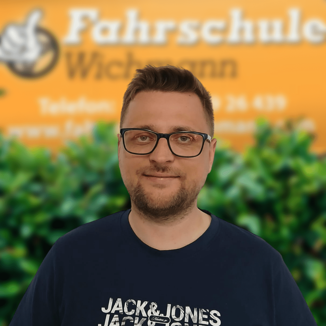 Mirco, Fahrlehrer der Fahrschule Wichmann in Delmenhorst, begleitet dich kompetent auf dem Weg zum Auto-, Motorrad-, LKW- oder Anhängerführerschein – auch in der Intensivfahrschule oder bei der Fahrlehrerausbildung. - Fahrschule Wichmann Delmenhorst - Autoführerschein LKW Führerschein Motorradführerschein Mirco, Fahrlehrer der Fahrschule Wichmann Delmenhorst, vor dem Firmenschild mit grünem Busch im Hintergrund.