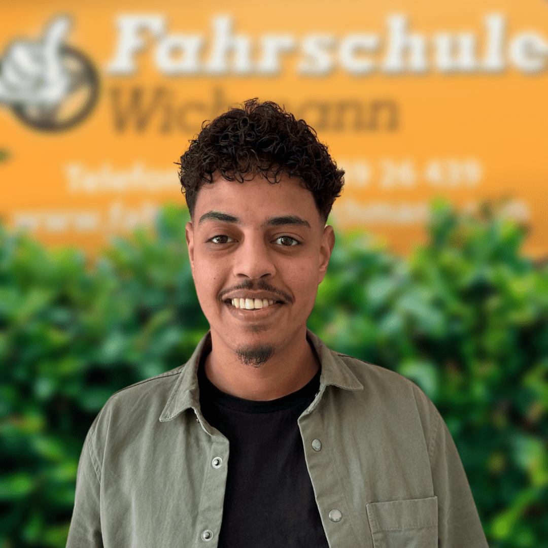 Fahrlehrer Sofiane von der Fahrschule Wichmann in Delmenhorst begleitet Dich kompetent beim Autoführerschein, Motorradführerschein, LKW- und Anhängerführerschein sowie in der Intensivfahrschule und Fahrlehrerausbildung. - Fahrschule Wichmann Delmenhorst - Autoführerschein LKW Führerschein Motorradführerschein Fahrlehrer Sofiane der Fahrschule Wichmann Delmenhorst vor grünem Busch und gelbem Firmenschild.