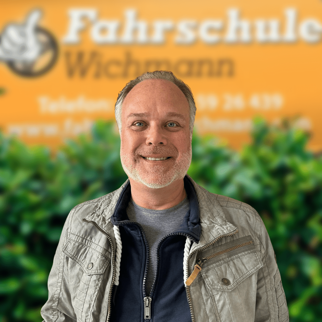 Sven, Fahrlehrer der Fahrschule Wichmann in Delmenhorst, begleitet dich kompetent zum Autoführerschein, Motorradführerschein, LKW- und Anhängerführerschein – auch in der Intensivfahrschule oder bei der Fahrlehrerausbildung. - Fahrschule Wichmann Delmenhorst - Autoführerschein LKW Führerschein Motorradführerschein Sven, Fahrlehrer der Fahrschule Wichmann in Delmenhorst, lächelt vor dem gelben Firmenschild mit grüner Hecke.