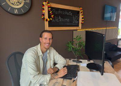 Man in a striped shirt sits at a wooden desk, typing on a keyboard with a monitor nearby in a brown-walled office. A chalkboard sign reading 'Fahrschule Wichmann' and garlands hang behind him.